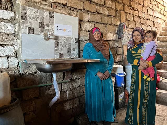Two women and a small child beside a newly installed handwashing basin in a brick home, with a Rotary plaque on the wall